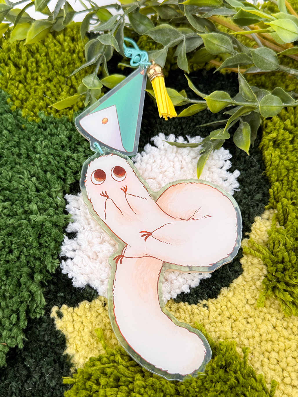 The Puffpuff ~ Acrylic Keychain features a cute, cartoonish white worm with big eyes, attached to a pastel geometric charm and yellow tassel, displayed on a green and yellow rug with artificial greenery.