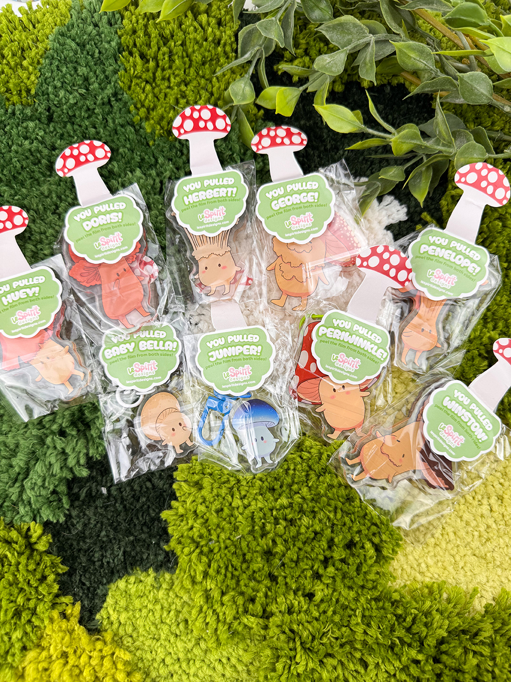 Mushroom Log ~ Mystery Gacha Mushroom Keychain Pulls in clear packaging, each with a cartoon mushroom tag and names like Doris, Herbert, Cooper, Cecilia, Baby Bella, and Juniper, are displayed on a leafy textured surface.