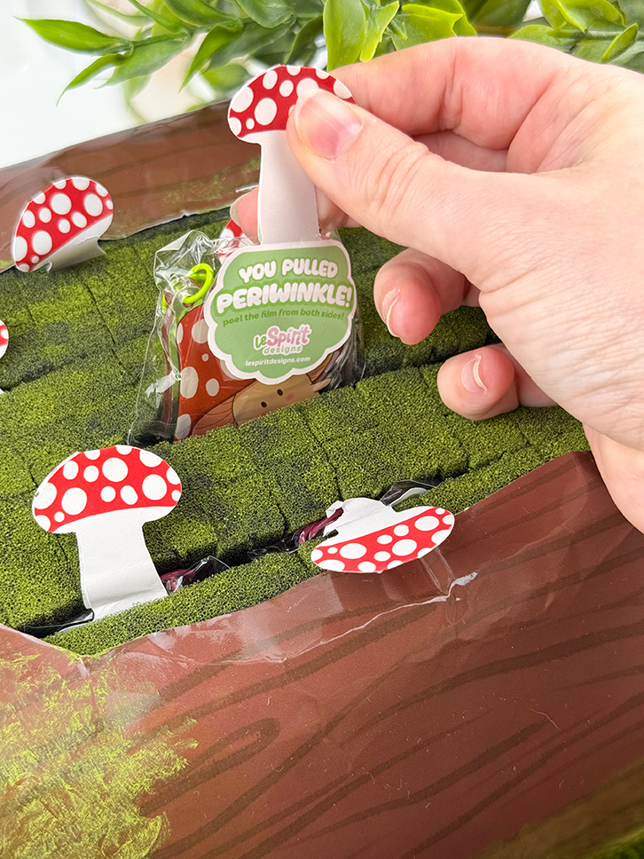 A hand pulls a red and white spotted Mushroom Log ~ Mystery Gacha Mushroom Keychain from a green mossy surface among similar mushroom tabs, with green leaves and a branded plastic bag in the background.
