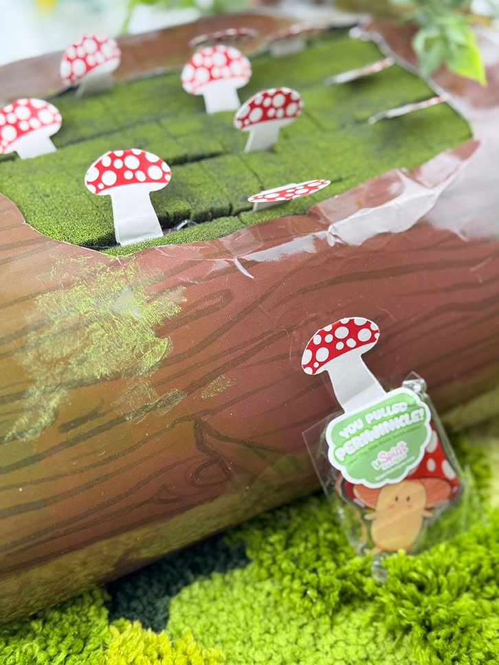 A close-up of the Mushroom Log ~ Mystery Gacha Mushroom Keychain Pulls game prop features red and white spotted mushroom pieces on green moss. One mushroom is pulled out, showing a prize tag that says, "You pulled a Shroomie!.