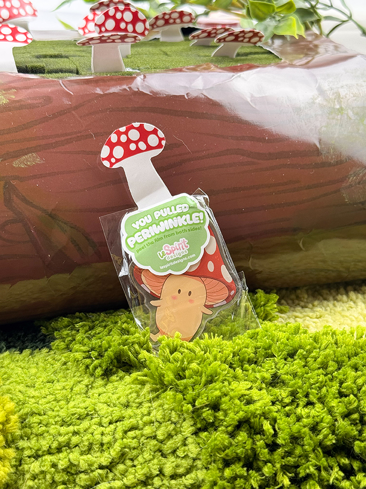 The Mushroom Log ~ Mystery Gacha Mushroom Keychain Pulls features a cute periwinkle mushroom character with a red and white cap on green grass, accented by tree log and mushroom decorations in the background.