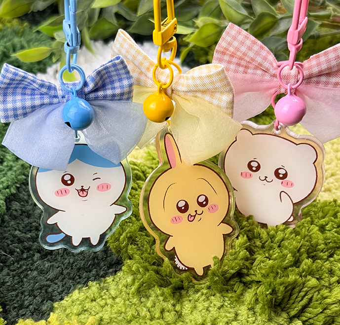 A cute Pink Hamster acrylic keychain hangs from a colorful clip with a bow and bell, set against green foliage and a textured rug.