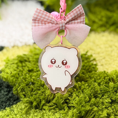 The Pink Hamster ~ Acrylic Keychain features a cute white cartoon hamster with blushing cheeks and big eyes, hanging below a pink and beige plaid bow, displayed on a green textured surface.