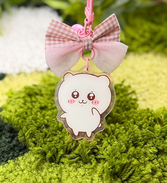The Pink Hamster ~ Acrylic Keychain features a cute white cartoon hamster with blushing cheeks and big eyes, hanging below a pink and beige plaid bow, displayed on a green textured surface.