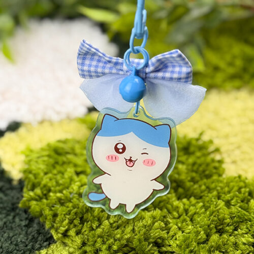 The Blue Kitty ~ Acrylic Keychain features a winking cartoon cat with blue hair, pink cheeks, a single fang, a blue bell, and a checkered bow, shown against a blurred green background.