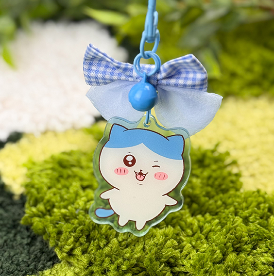 The Blue Kitty ~ Acrylic Keychain features a winking cartoon cat with blue hair, pink cheeks, a single fang, a blue bell, and a checkered bow, shown against a blurred green background.