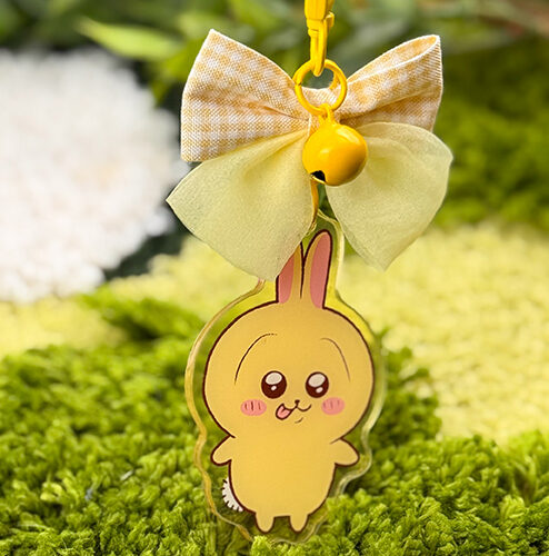 The Yellow Bunny ~ Acrylic Keychain features a cute yellow bunny with pink cheeks and ears, a yellow bow, and a small bell, displayed above green fuzzy grass with blurred foliage in the background.