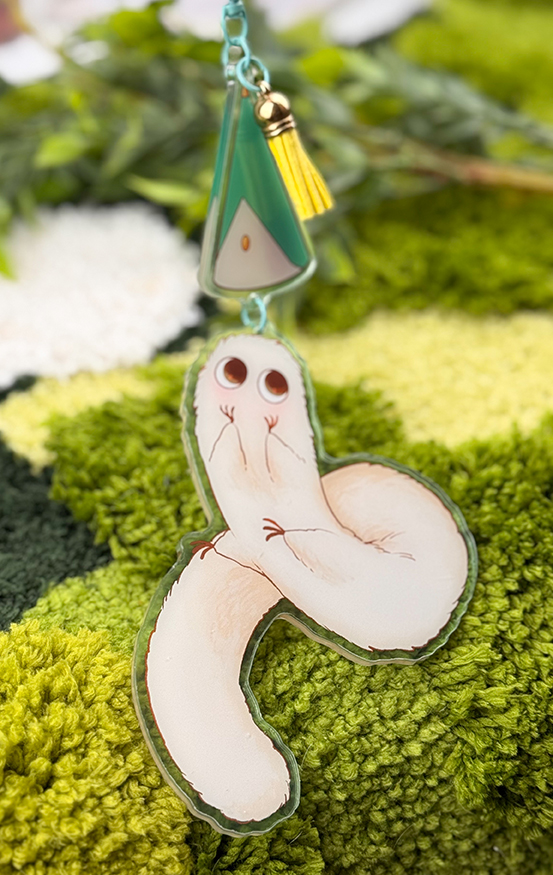 The Puffpuff ~ Acrylic Keychain features a quirky, cartoonish white creature with big eyes and a triangular charm, shown on a fuzzy green surface with foliage in the background.
