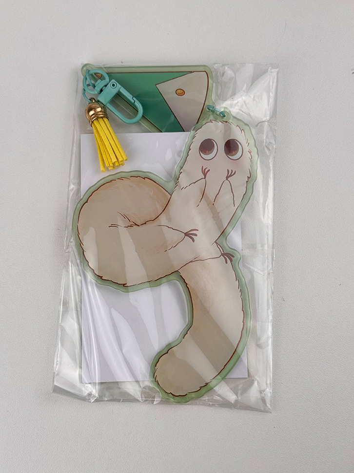 The Puffpuff ~ Acrylic Keychain is a clear plastic keychain featuring a cute cartoon animal with big eyes and a fluffy curled tail, attached to a green and yellow tassel charm, and comes on a white paper backing.