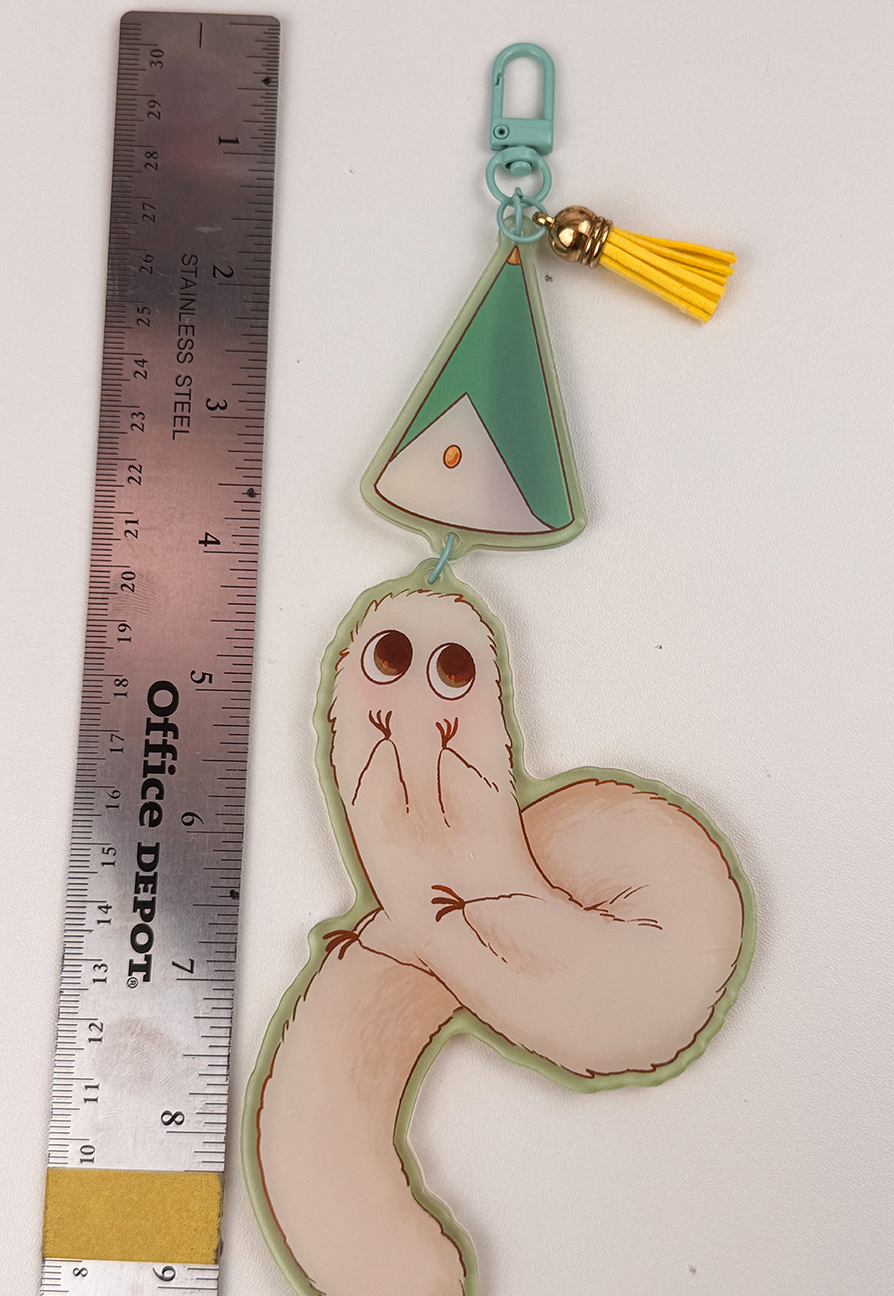 The Puffpuff ~ Acrylic Keychain features a cartoonish, pale, worm-like creature, a green triangle charm, and a yellow tassel. Shown next to a 6-inch ruler for scale, the keychain measures about 7 inches long.