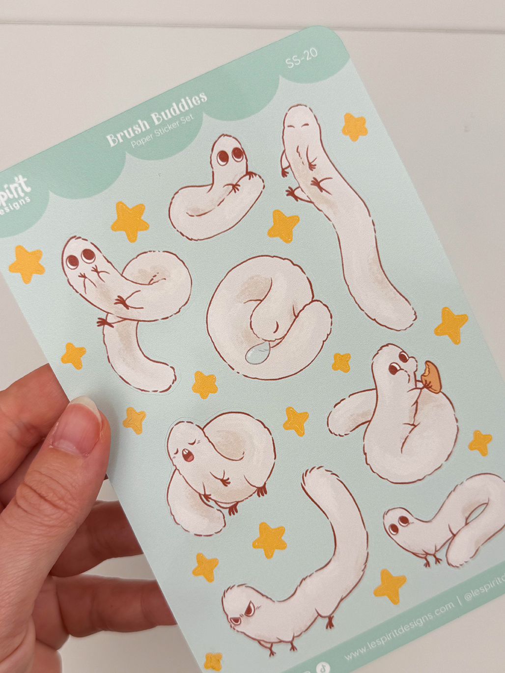 A hand holds the Puffpuff ~ Paper Sticker Sheet, showcasing adorable, fluffy white ferret-like creatures in playful poses, surrounded by yellow stars.