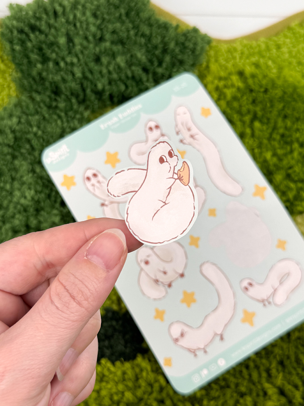 A hand holds a sticker from the Puffpuff ~ Paper Sticker Sheet, featuring a ghost-like creature eating a cookie, with more whimsical ghosts and yellow stars on the sheet in the background, set against a green textured surface.