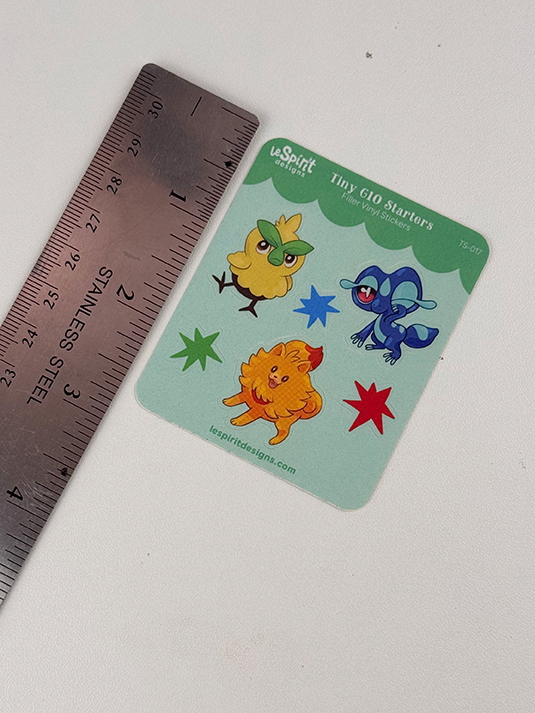 The Tiny Gen10 Starters ~ Tiny Filler Vinyl Sticker Sheet features cartoon animals like a yellow chick, blue lizard, orange puppy, and blue frog among colorful stars on green, shown next to a stainless steel ruler.