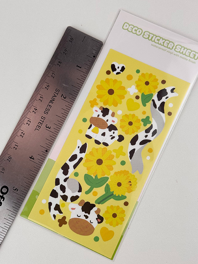 The Sunflower Cows ~ Deco Sticker Sheet features cartoon cows, yellow sunflowers, and hearts on a yellow background. A stainless steel ruler is shown next to the sheet for size reference.