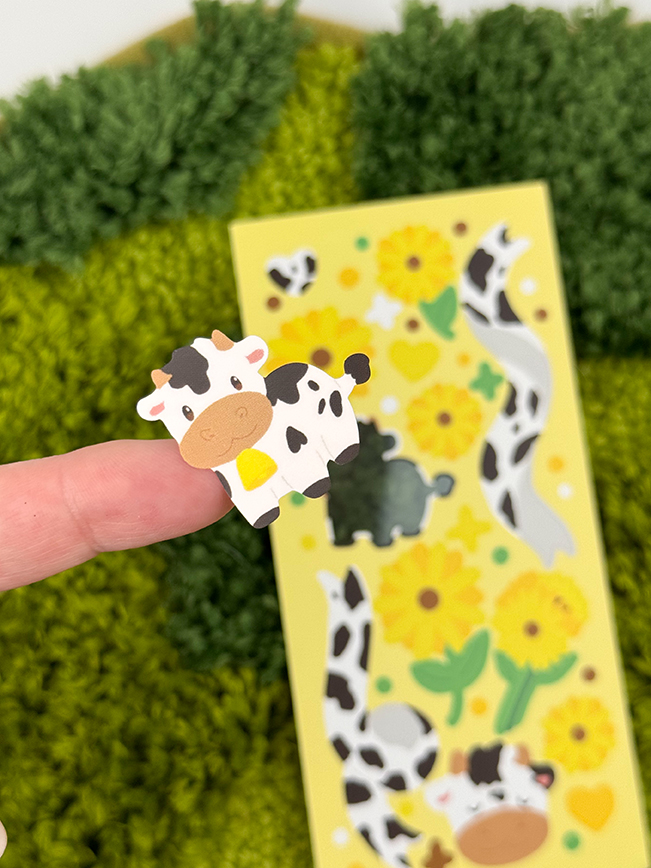 A finger holds a cute cartoon cow sticker from the Sunflower Cows ~ Deco Sticker Sheet. The yellow sheet in the background features sunflowers, hearts, and other cow-themed stickers on a green textured surface.