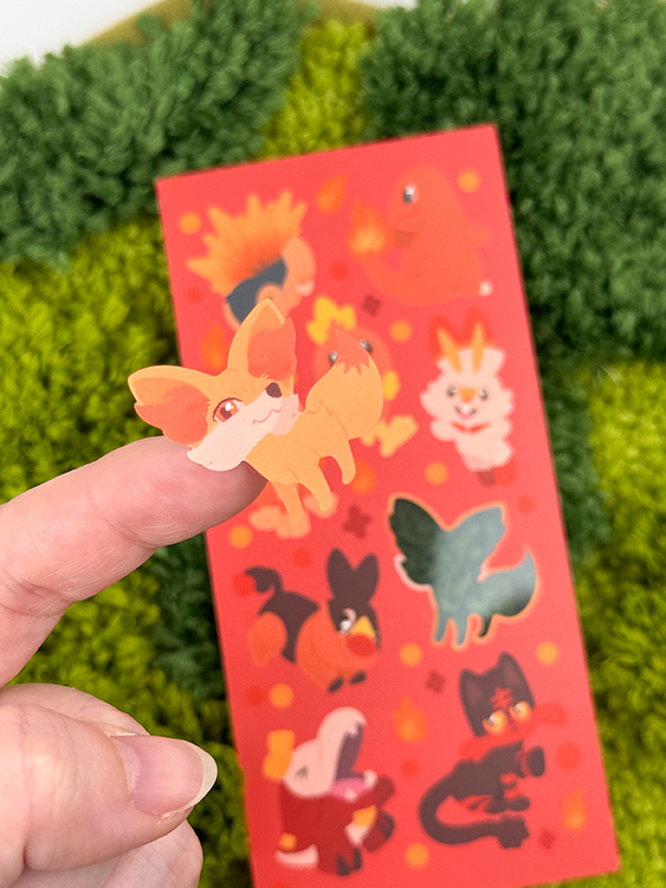 A hand displays a small sticker of an orange fox from the Fire Starters ~ Deco Sticker Sheet, held over a red sheet with colorful animal stickers on a green textured surface.