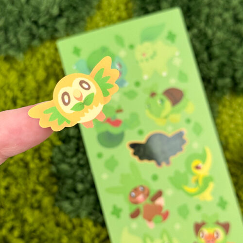 A close-up of a finger holding a round, green bird sticker from the Grass Starters ~ Deco Sticker Sheet, featuring cute green animal characters on a leafy background.