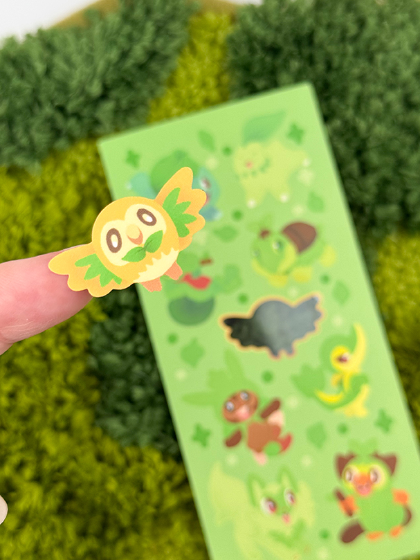 A close-up of a finger holding a round, green bird sticker from the Grass Starters ~ Deco Sticker Sheet, featuring cute green animal characters on a leafy background.