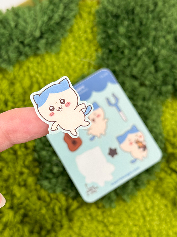 A hand holds a "Tiny Blue Kitty ~ Tiny Filler Vinyl Sticker Sheet" sticker featuring a cartoon cat with a blue cap. More similar cat stickers are seen on a light blue sheet in the background, set against green grass.