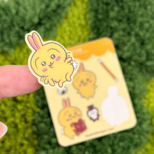 A hand holds a cute sticker from the "Tiny Yellow Bunny ~ Tiny Filler Vinyl Sticker Sheet," featuring a yellow bunny with pink cheeks and ears. In the background, the matching sticker sheet lies on a green textured surface.