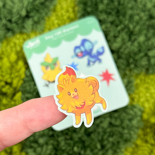 A hand holds a small, colorful lion sticker from the Tiny Gen10 Starters ~ Tiny Filler Vinyl Sticker Sheet. In the blurred background, a card shows more cartoon animal stickers and a red star on green textured fabric.