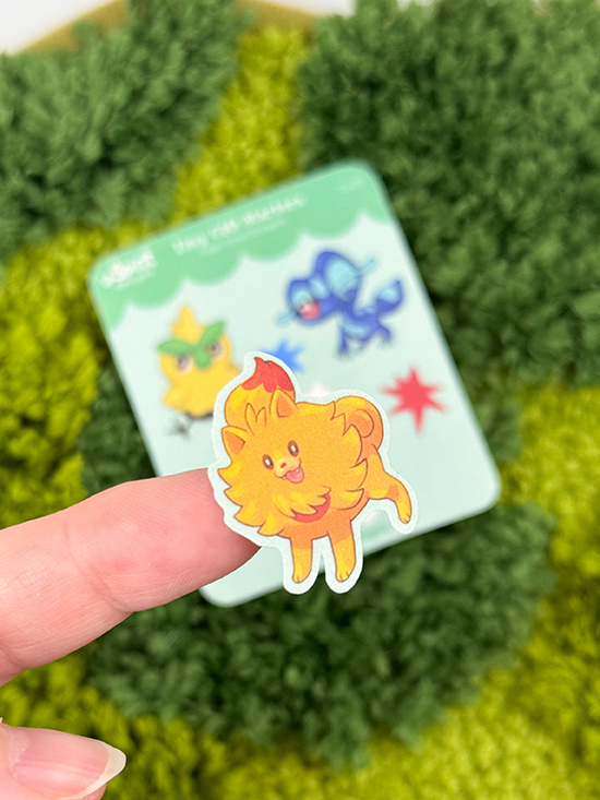 A hand holds a small, colorful lion sticker from the Tiny Gen10 Starters ~ Tiny Filler Vinyl Sticker Sheet. In the blurred background, a card shows more cartoon animal stickers and a red star on green textured fabric.