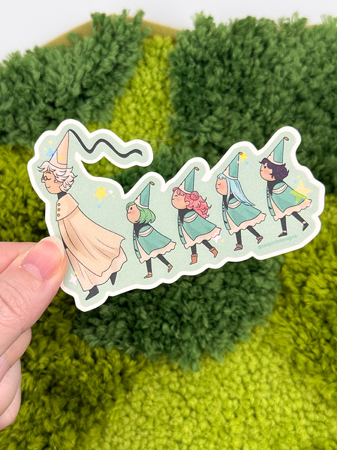 A hand holds the Uppies Kitty ~ 3" Matte Vinyl Sticker, featuring five whimsical characters in wizard hats and cloaks walking in a line against a green, textured, fuzzy background.