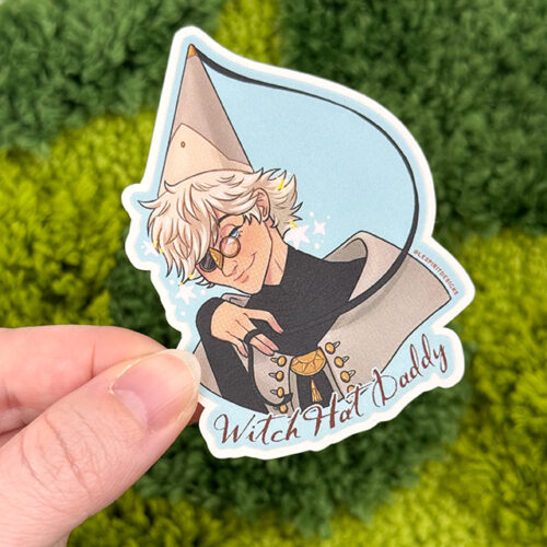 A hand holds the Uppies Kitty ~ 3" Matte Vinyl Sticker, featuring an illustrated cat character with a witch hat and glasses, smiling mischievously against a green mossy background.