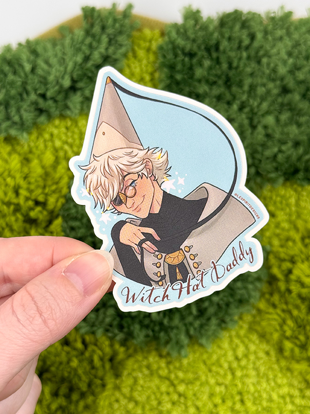 A hand holds the Uppies Kitty ~ 3" Matte Vinyl Sticker, featuring an illustrated cat character with a witch hat and glasses, smiling mischievously against a green mossy background.