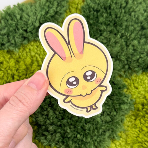 A hand holds the Uppies Kitty ~ 3" Matte Vinyl Sticker, featuring an adorable yellow cartoon bunny with pink ears, big sparkling eyes, and blushing cheeks, set against a green textured background.