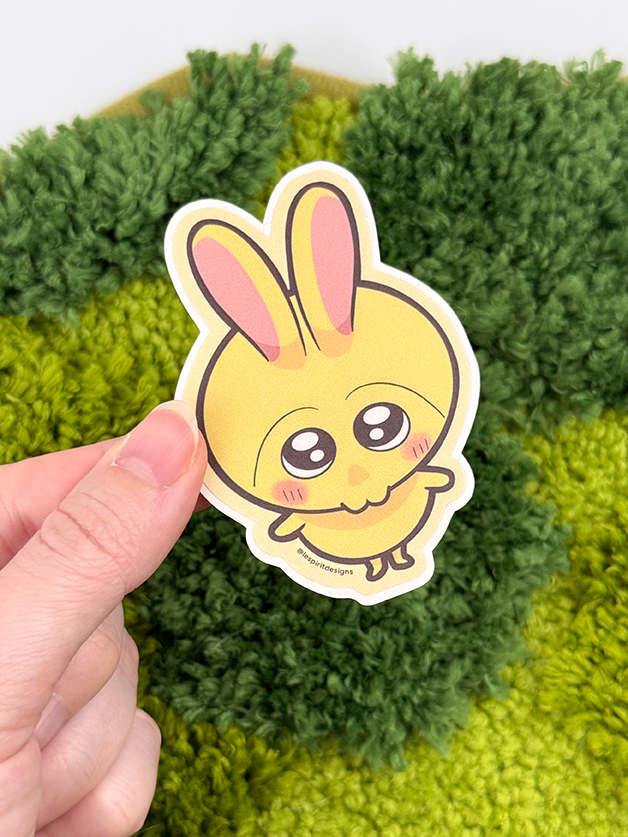 A hand holds the Uppies Kitty ~ 3" Matte Vinyl Sticker, featuring an adorable yellow cartoon bunny with pink ears, big sparkling eyes, and blushing cheeks, set against a green textured background.
