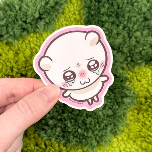 A hand holds the Uppies Hamster ~ 3" Matte Vinyl Sticker, featuring a cute cartoon hamster with blushing cheeks and big teary eyes against a textured green grassy background.