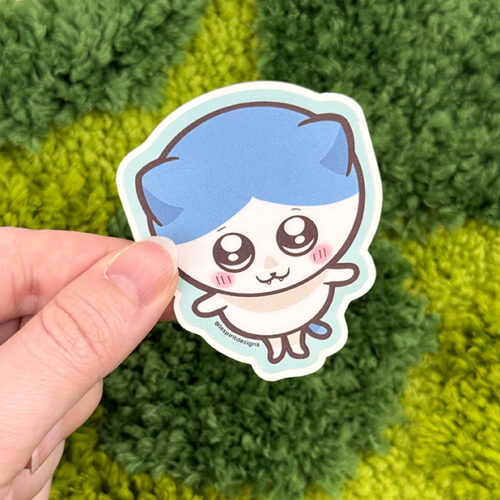 A hand holds the Uppies Kitty ~ 3" Matte Vinyl Sticker, featuring a cute cartoon kitty with blue hair, big eyes, blushing cheeks, and tiny limbs. The sticker is displayed against textured green fabric resembling grass.