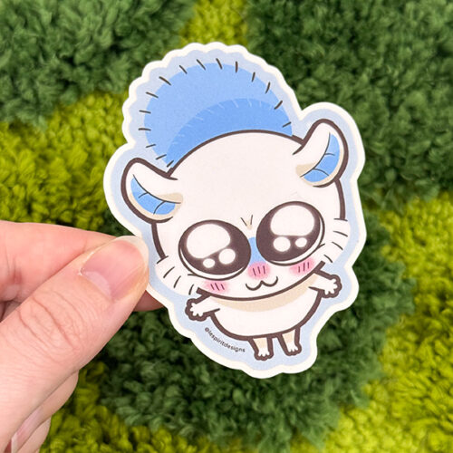 A hand holds the Uppies Kitty ~ 3" Matte Vinyl Sticker, featuring a cute, wide-eyed white cat with blushing cheeks and small paws. The background is a textured, green, grass-like surface.