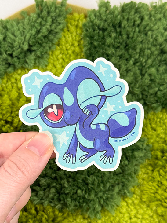 A hand holds the Uppies Kitty ~ 3" Matte Vinyl Sticker, showing a cute, cartoon-style blue gecko-like creature with large eyes and star accents on a fuzzy green and yellow background.