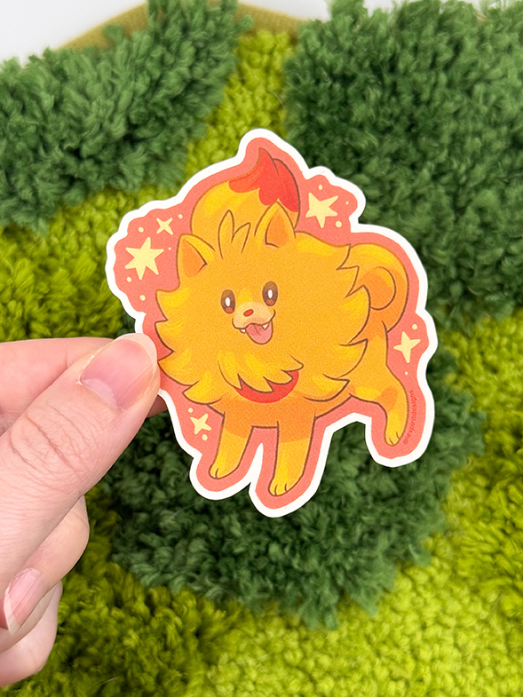 A hand holds the Uppies Kitty ~ 3" Matte Vinyl Sticker, featuring a cute cartoon cat with a fluffy tail and cheerful expression, surrounded by stars against fuzzy green grass-like fabric.