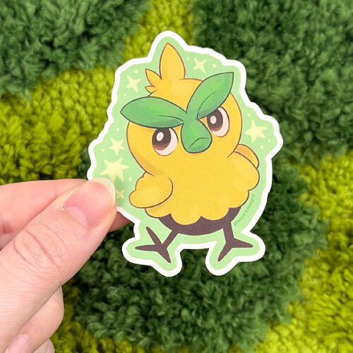 A hand holds the Uppies Kitty ~ 3" Matte Vinyl Sticker, featuring a cute, cartoon yellow bird with green leaf eyebrows on a green starry background. The backdrop is textured green fabric.