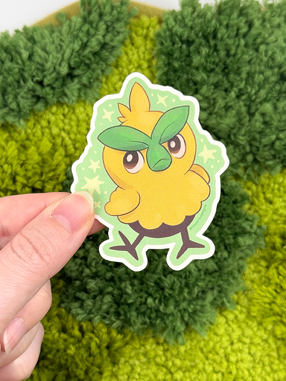 A hand holds the Uppies Kitty ~ 3" Matte Vinyl Sticker, featuring a cute, cartoon yellow bird with green leaf eyebrows on a green starry background. The backdrop is textured green fabric.