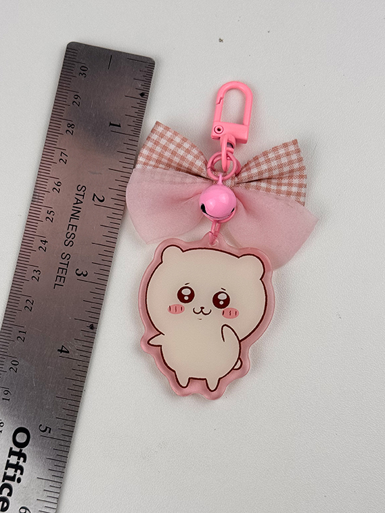 The Pink Hamster ~ Acrylic Keychain features a cute, blushing cartoon hamster, a pink bow, and a small bell. It has a pink clip and is shown beside a stainless steel ruler for scale.