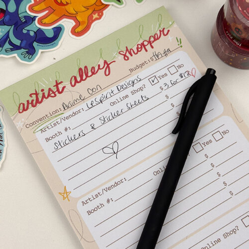 The Artist Alley Shopper ~ 50 page Notepad features handwritten notes about buying stickers from LeftSpirit Designs at Anime Con, with a black pen on top and surrounded by colorful stickers and washi tape rolls.