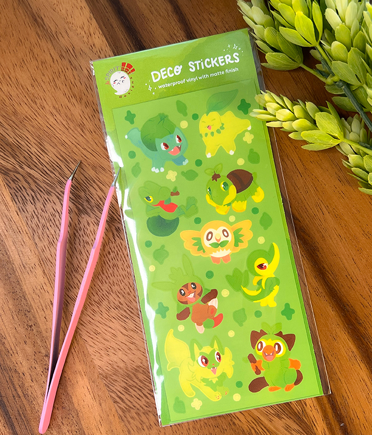 A sheet of "Grass Starters ~ Deco Sticker Sheet" with cute cartoon animal designs is shown on a wooden surface beside two pink tweezers and artificial green foliage.