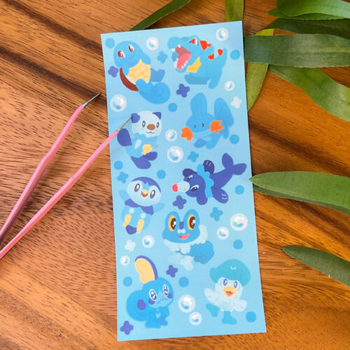 A Water Starters ~ Deco Sticker Sheet featuring cute, colorful water-type Pokémon on a blue background with bubble and star designs, shown on a wooden surface beside a green plant and pink tweezers.