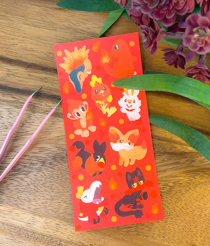 The Fire Starters ~ Deco Sticker Sheet showcases cartoon fire-themed Pokémon with green leaves and a purple-flowered plant, displayed on wood with pink tweezers nearby.
