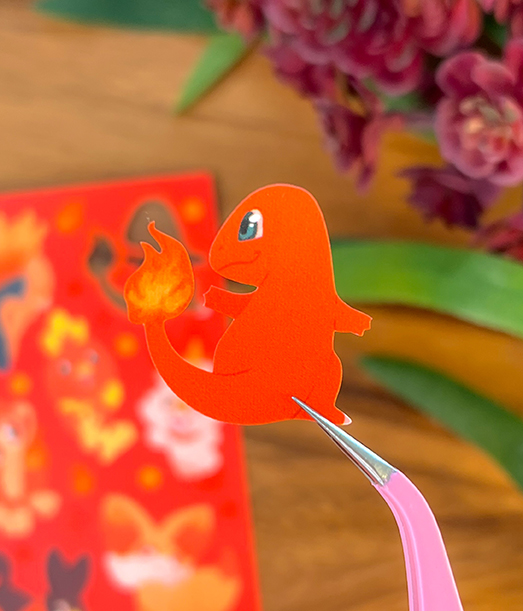 A hand holds pink tweezers to lift a Charmander sticker from the Fire Starters ~ Deco Sticker Sheet, with a blurred floral background and other red stickers visible.