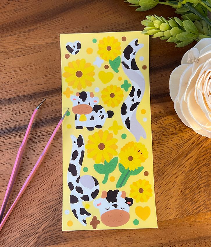 The Sunflower Cows ~ Deco Sticker Sheet is displayed on a wooden surface beside two pink tools and artificial white and green flowers.