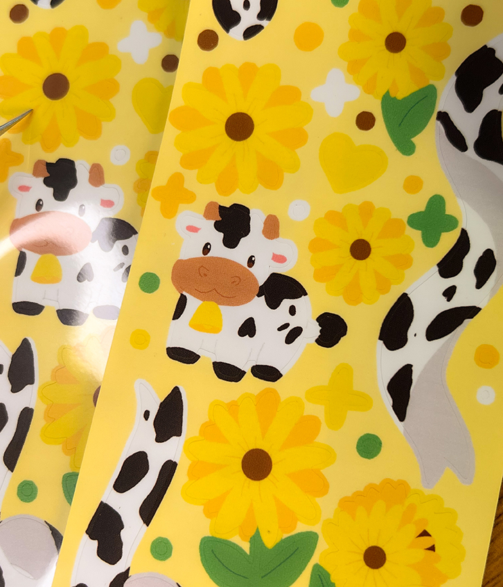 The Sunflower Cows ~ Deco Sticker Sheet features cartoon cows, yellow flowers, green leaves, and cow print patterns on a bright yellow background.