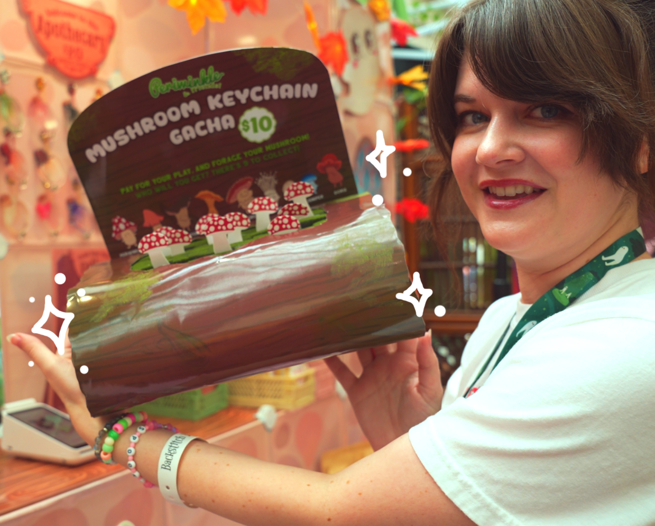 A smiling person holds a display box labeled "Mushroom Log ~ Mystery Gacha Mushroom Keychain Pulls" ($10) with small mushroom keychains inside. Decorative sparkles and a colorful, whimsical background complete the scene.