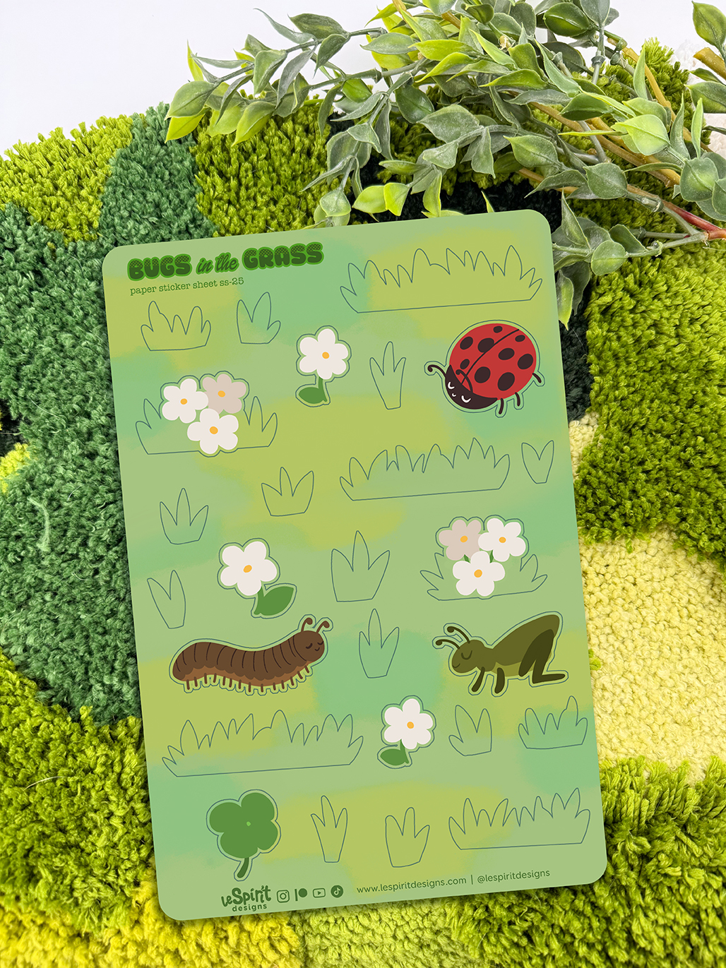 Bugs in the Grass ~ Paper Sticker Sheet features cartoon bugs like a ladybug, centipede, and grasshopper, along with flowers and grass illustrations on textured green paper, with a leafy plant in the background.