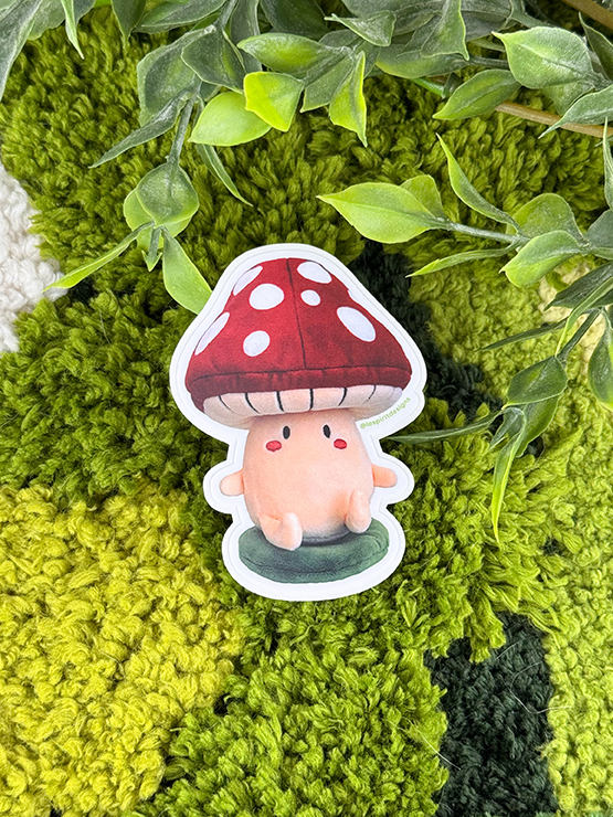 A cute cartoon mushroom character sticker sits on green textured yarn with leafy plant stems above, creating a playful forest vibe.
