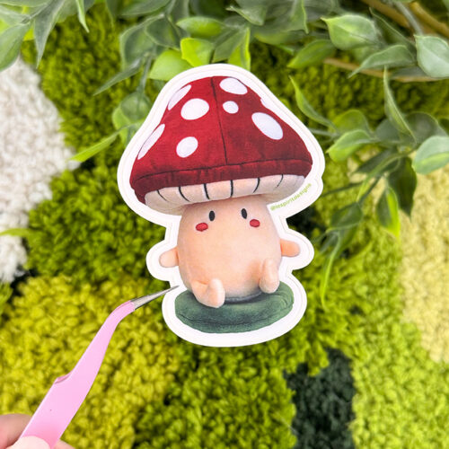 A hand holds tweezers gripping a cute mushroom sticker with a red cap and white spots, against a background of green plants and textured surface.